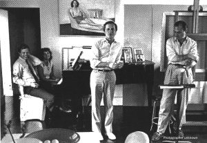 Paul Kellogg leans on a piano. There is a painting of a woman on the wall behind him. To his right and left are men. A woman sits at the piano