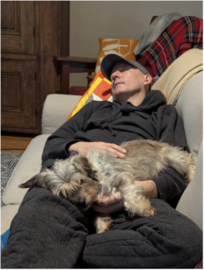 Rob Ainsley lying on a couch holding a dog.
